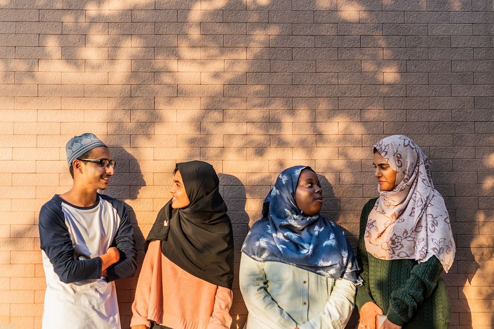 Group of four muslim teenagers standing against a brick wall, wearing hijabs and casual clothing, enjoying a sunny day.…