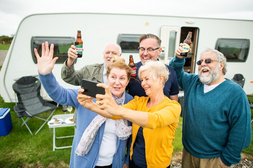 Group of older adults enjoying a day outside a camper, holding drinks and taking a selfie. Smiling and cheerful, they…