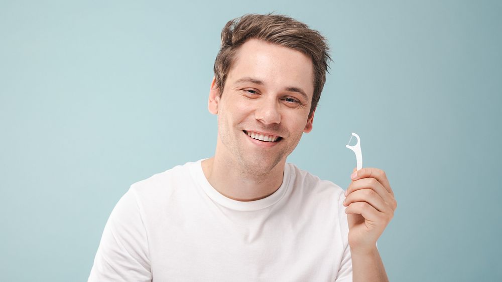 Smiling man holding a dental floss pick against a blue background. Dental care, flossing, and oral hygiene are key. Happy…