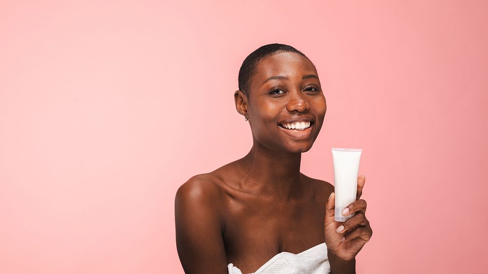 Smiling woman holding skincare product, pink background. Skincare focus with woman displaying product. Happy woman promoting…