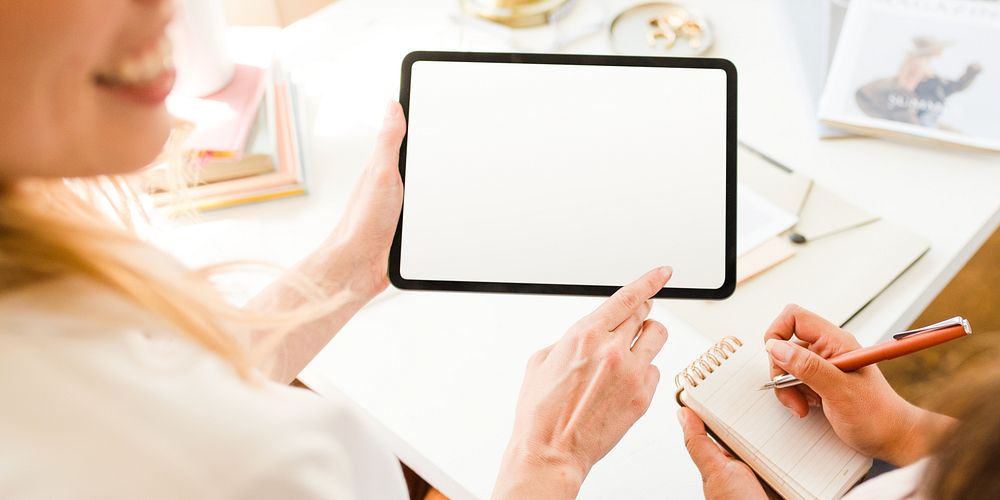 Two people collaborating, using a tablet and notebook. Bright workspace and technology. Hands writing, hands holding tablet…