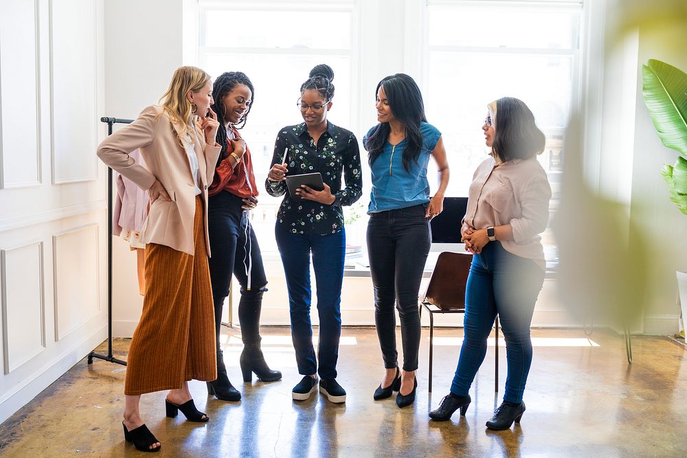 A diverse group of women in a bright office, discussing work. Diverse women standing together, smiling, and using a tablet.…