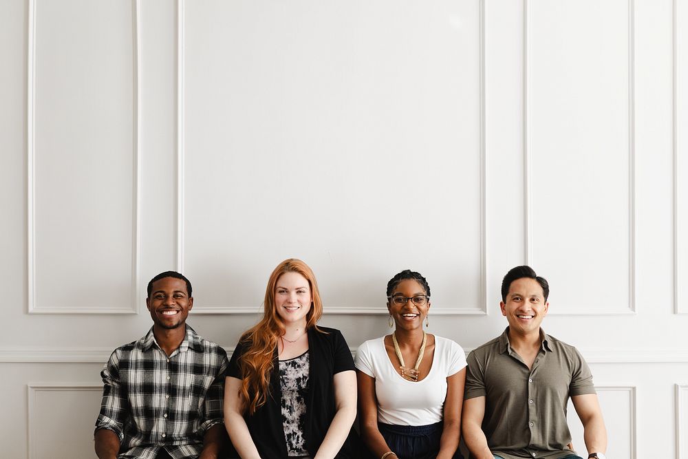 Group of four people sitting against a white wall, smiling. Diverse group includes men and women, showcasing diversity and…