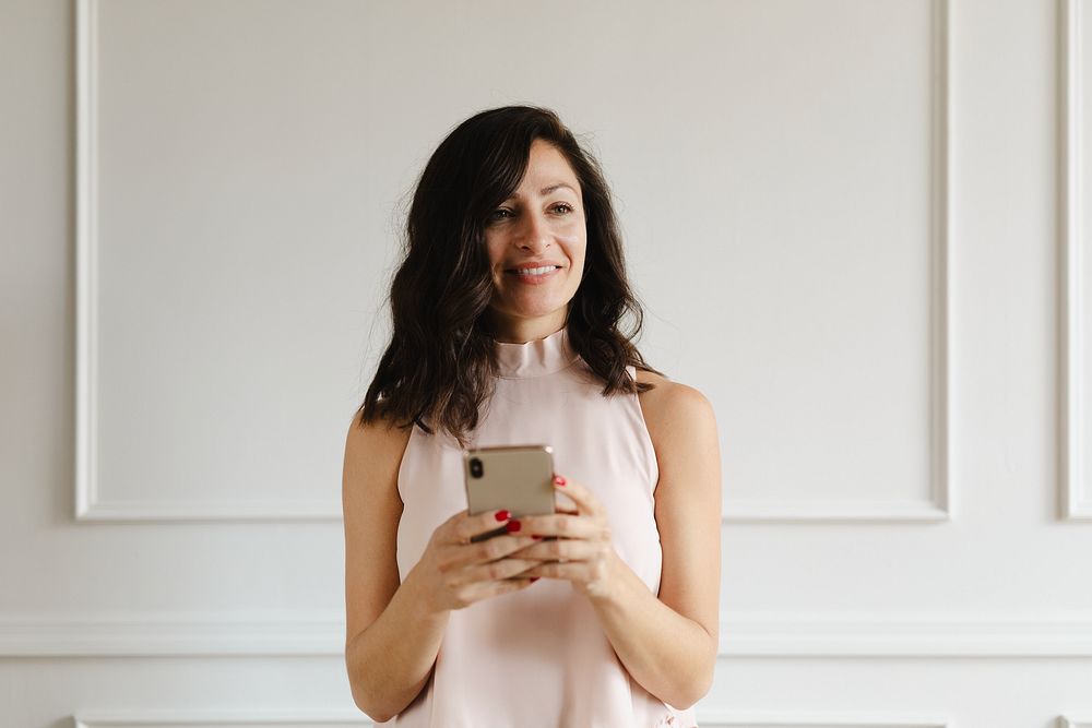 Smiling woman holding a smartphone, standing against a white wall. Casual attire, relaxed pose, smartphone in hand, modern…