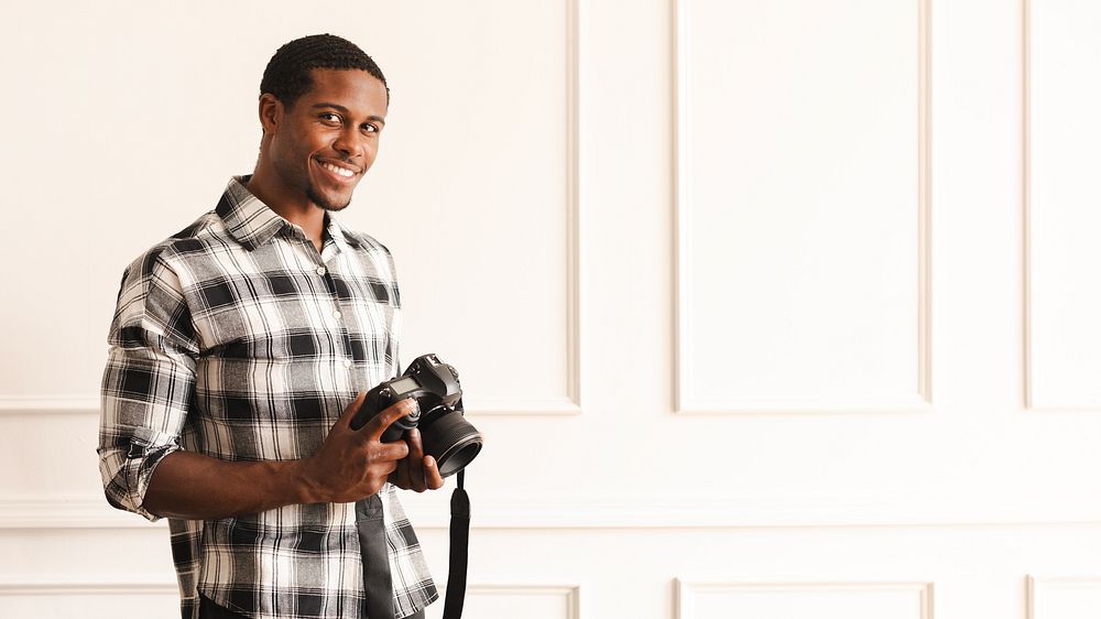 Man holding a camera, wearing a plaid shirt, smiling. Photography enthusiast, casual style, indoor setting. Capturing…