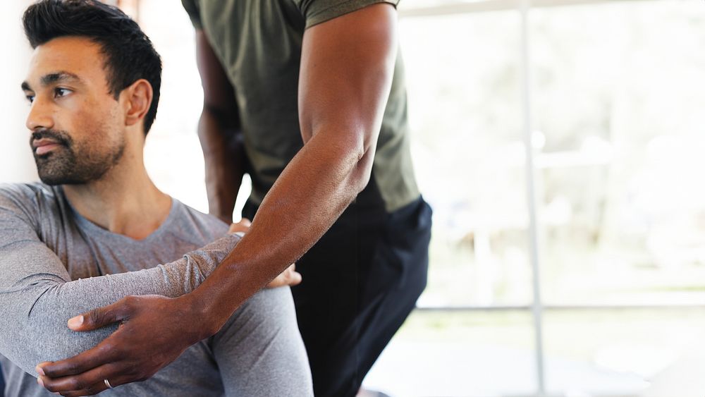 A person assists another in a stretching exercise. Focus on arm movement and physical support. Indoor setting with natural…