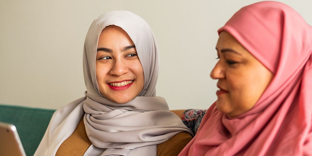 Two women in hijabs smiling, one in gray, one in pink. Women are sitting together, sharing a moment. Hijabs, smiles, and…