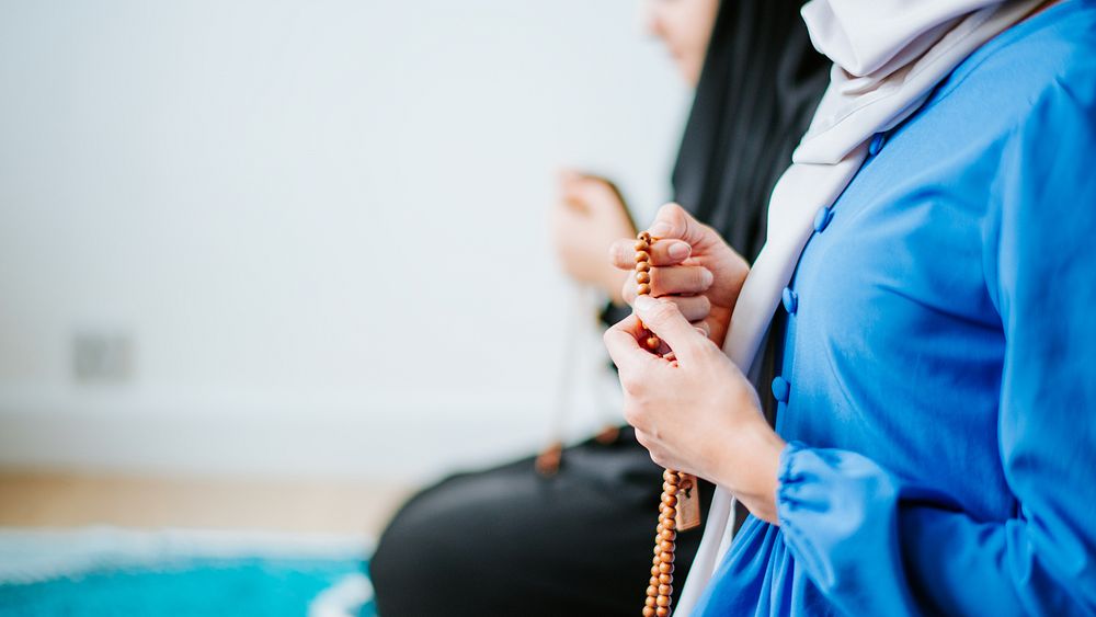 Two muslim women in traditional attire, one in blue, holding prayer beads. Focus on hands and beads, symbolizing prayer and…