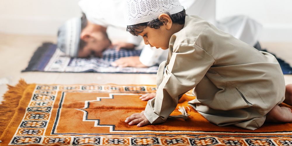 A young Arab boy in traditional attire prays on a prayer mat, showcasing devotion. The boy's focus on prayer highlights the…