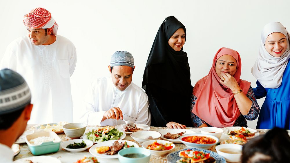 Muslim family and friends enjoying a meal with diverse dishes. Men and women in traditional attire. Smiling, sharing, and…