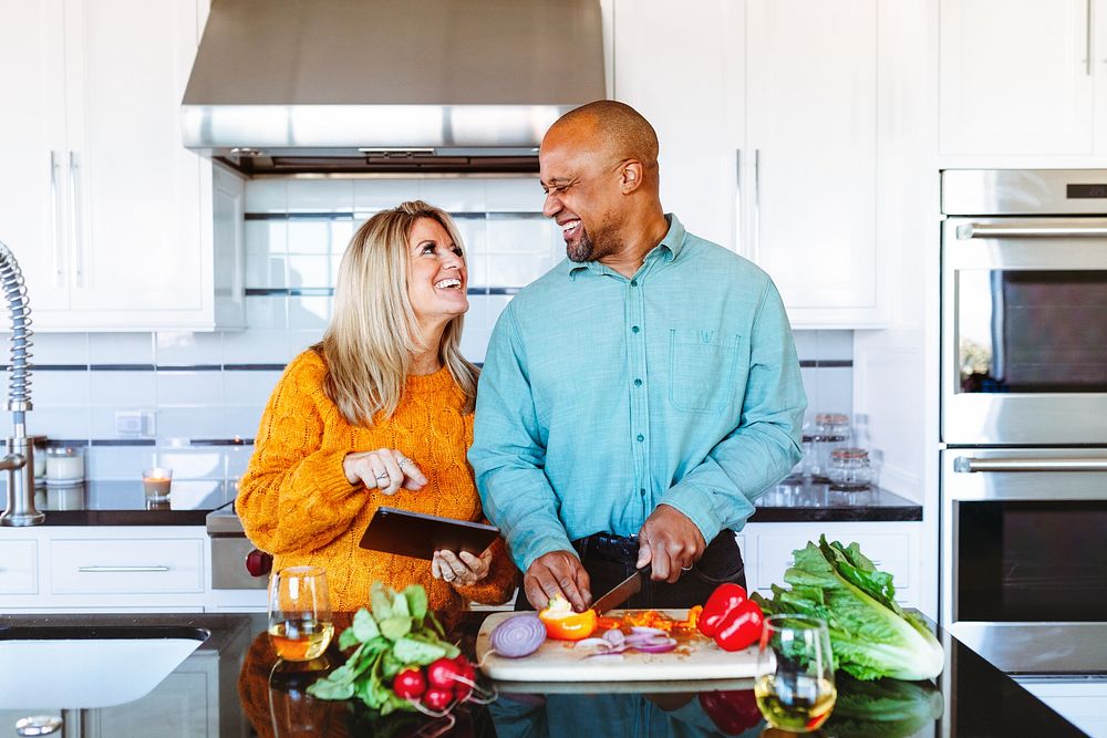 A happy couple, a Caucasian woman and an African American man, enjoy cooking together in a modern kitchen. They share smiles…