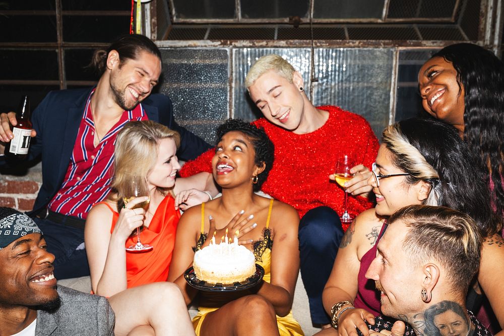 Group of diverse friends celebrating a birthday with cake and drinks, smiling and laughing together at a lively party.…