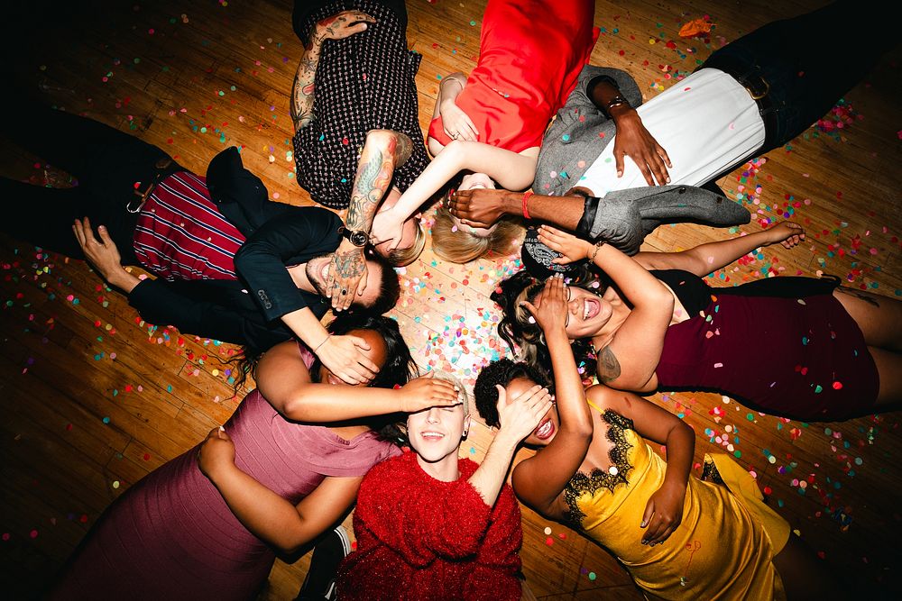 Diverse group of people lying on the floor, covering their eyes, surrounded by confetti. Celebration, joy, and diversity in…