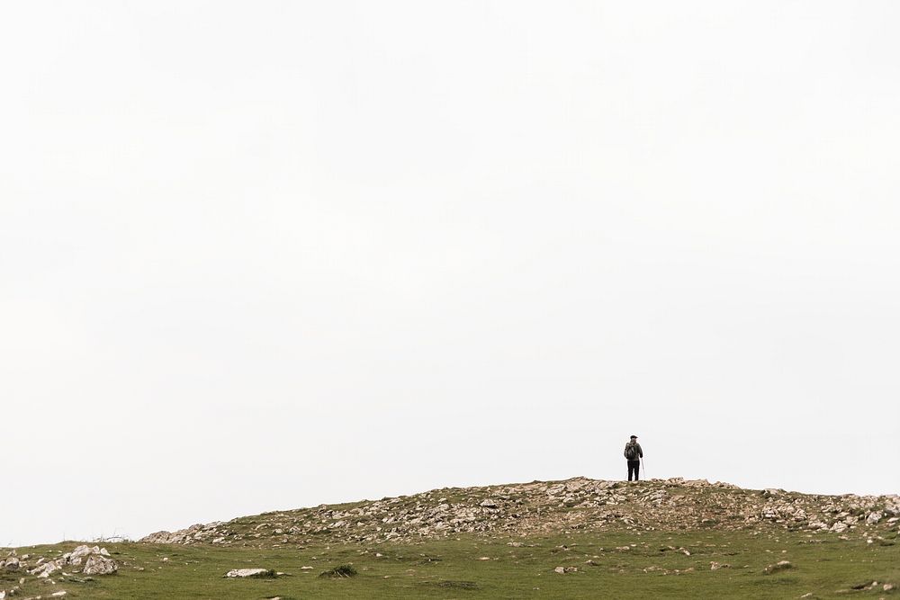 A solitary figure stands on a rocky hilltop under a vast, cloudy sky. The lone person on the hill top gazes at the expansive…