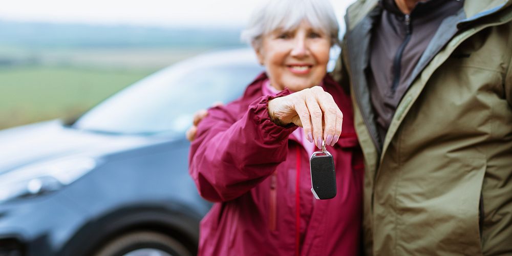 Elderly couple holding car keys, standing by a car. Outdoors, countryside. Senior man and woman, happy with new car. Car…