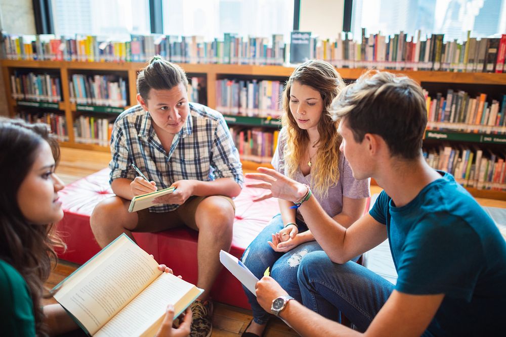 Group of young adults in a library, discussing books. Diverse group, engaged in conversation, surrounded by shelves of…