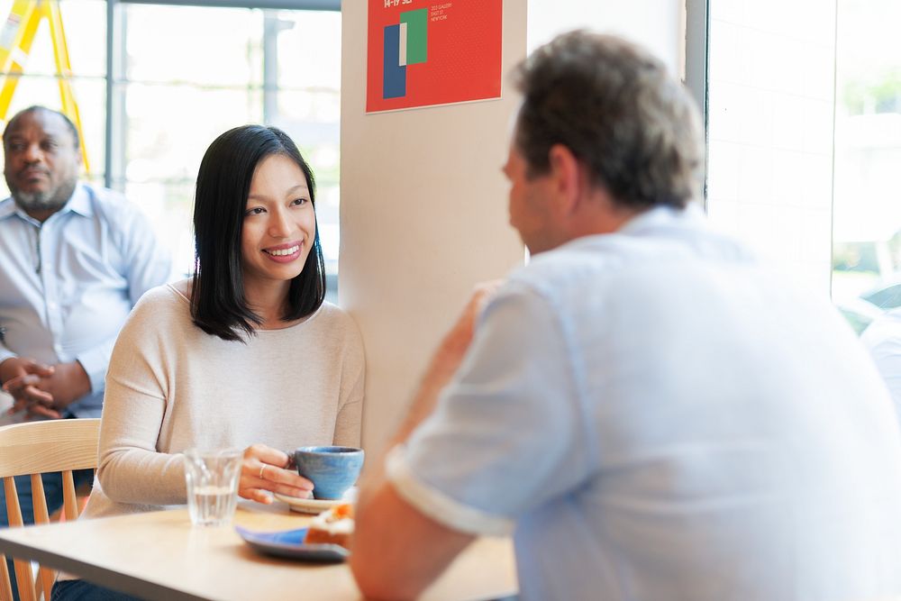 Two people at a cafe table, smiling and talking. Woman with long black hair, man with short hair. Casual setting, coffee…