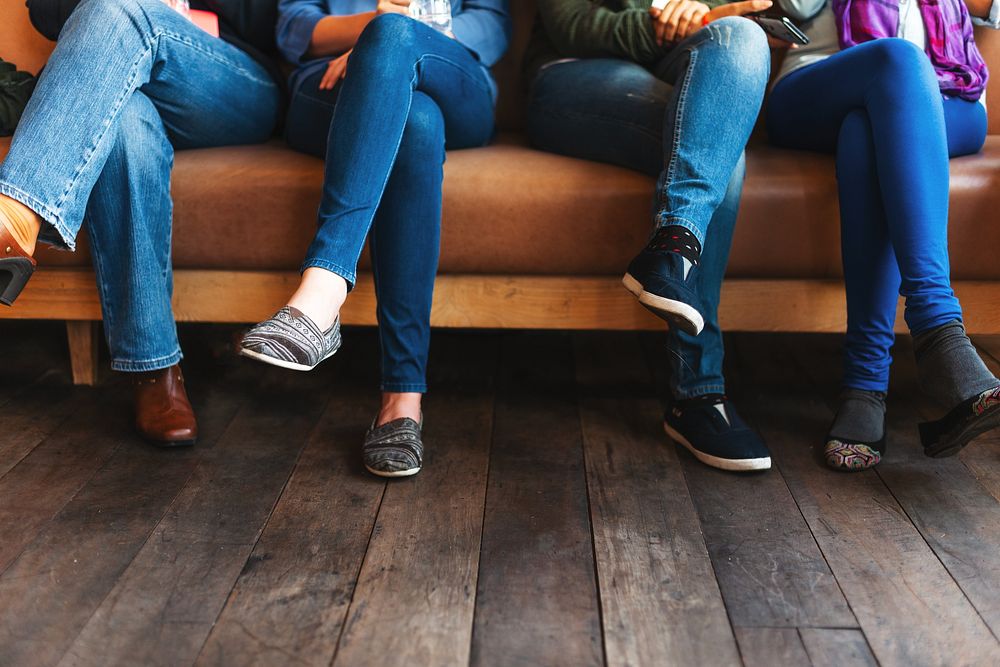 Four people sitting on a couch, legs crossed, wearing blue jeans and casual shoes. Wooden floor, casual setting, diverse…