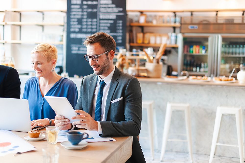 Two business people in a cafe, one using a tablet, the other a laptop. Bright modern setting with a casual business vibe.…