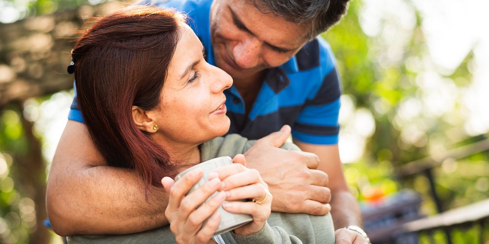 A couple enjoying a moment outdoors. The man embraces the woman lovingly. They share a warm connection, smiling and relaxed…