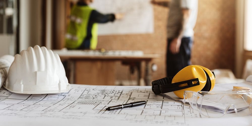 Construction site with blueprints, hard hat, and safety gear. Focus on planning and safety. Workers in background, blurred…