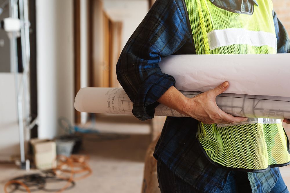 Person in construction gear holding blueprints. Construction worker with plans. Safety vest, blueprints, construction site.…