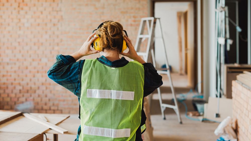 Female construction worker wearing safety gear, including a vest and headphones, in a building site. Worker focuses on…