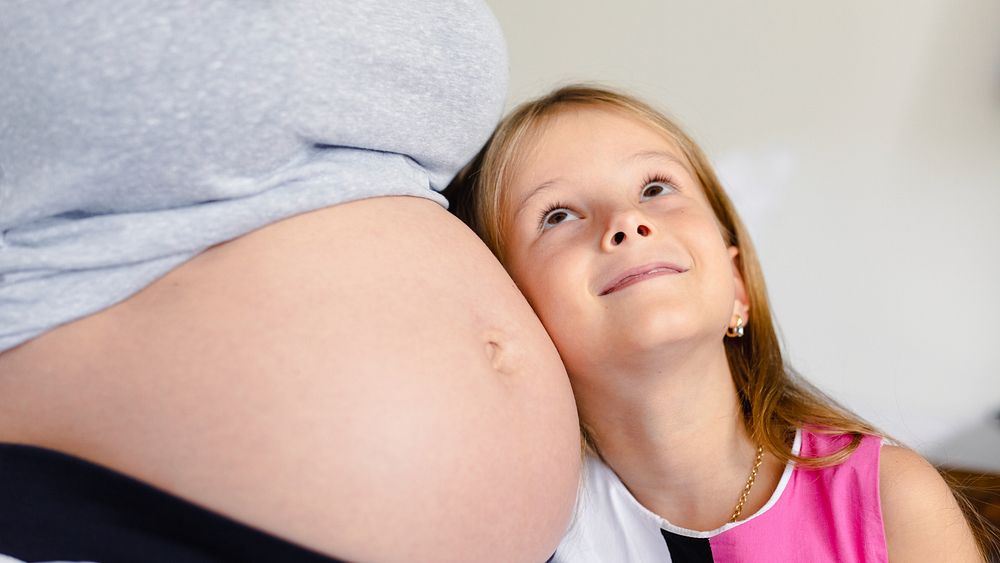 A young girl smiles, resting her head on a pregnant belly. The scene captures a tender moment of family, pregnancy, and…