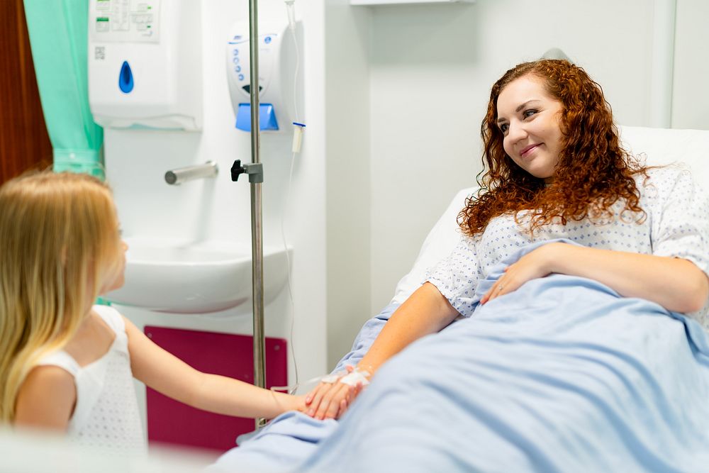 A woman with curly hair lies in a hospital bed, smiling at a young girl. Hospital setting, patient care, and comforting…