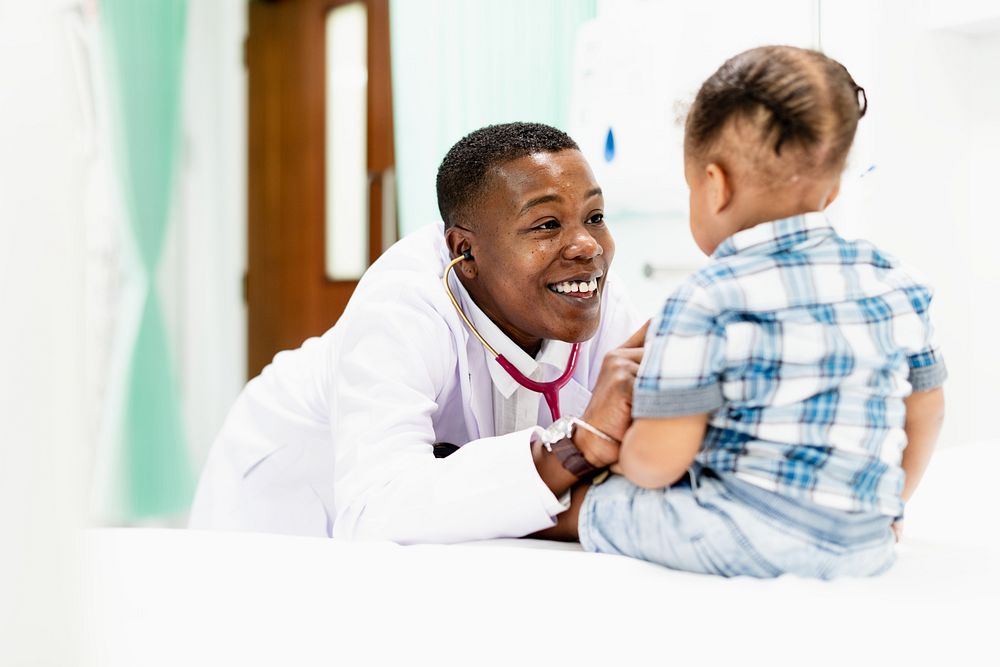 Doctor interacting with a child in a hospital setting. The doctor is smiling and wearing a white coat. The child is sitting…