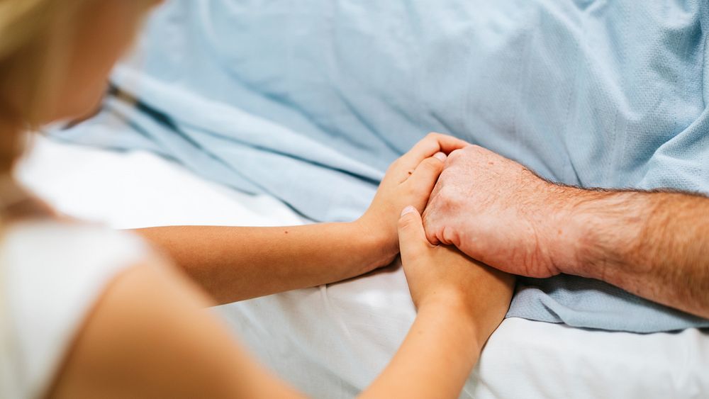 Close-up of two people holding hands, one lying in bed. Comforting hands, support, and care are shown. Hands clasped…