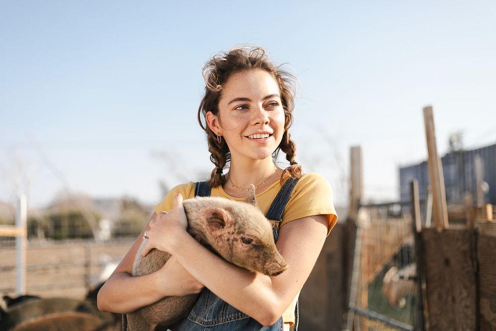 Young woman holding a piglet on a farm. Smiling woman with piglet, enjoying farm life. Farm setting with woman and piglet…