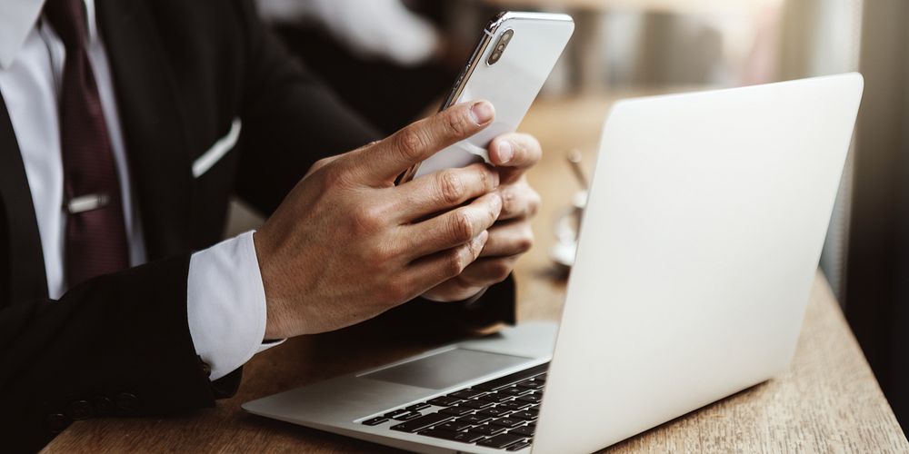 Person using smartphone and laptop at a desk. Hands holding phone, laptop open. Technology use, multitasking. Office…