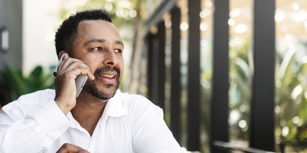 Man talking on phone outdoors, wearing a white shirt. Relaxed conversation, enjoying a sunny day. Greenery in the…