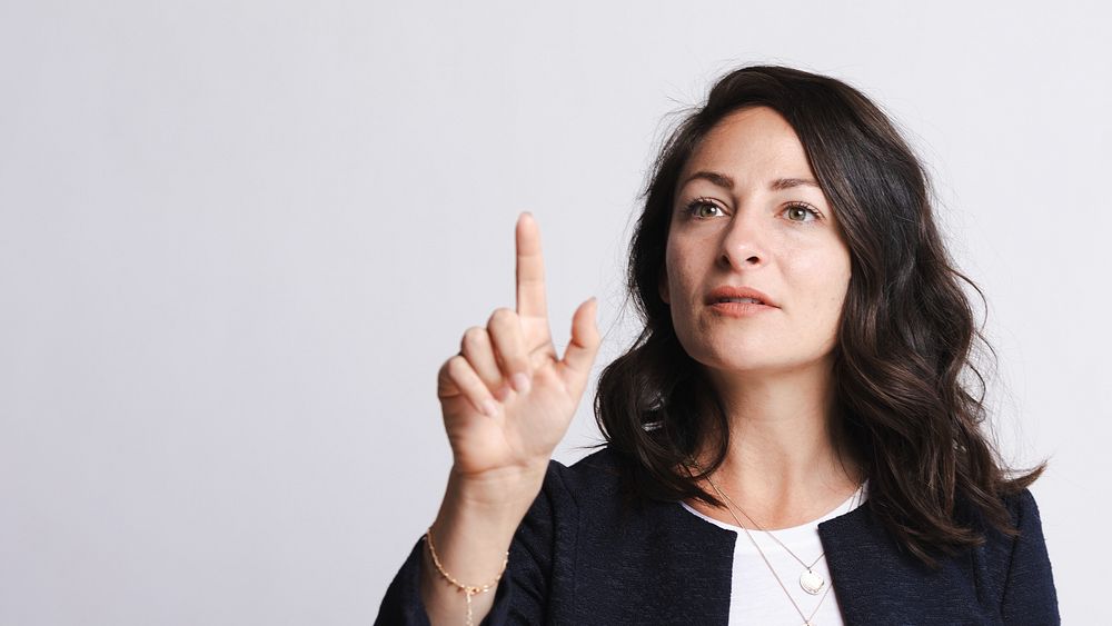 Woman gesturing with finger, focused expression. Brunette woman, pointing, wearing a dark blazer. Confident woman, making a…