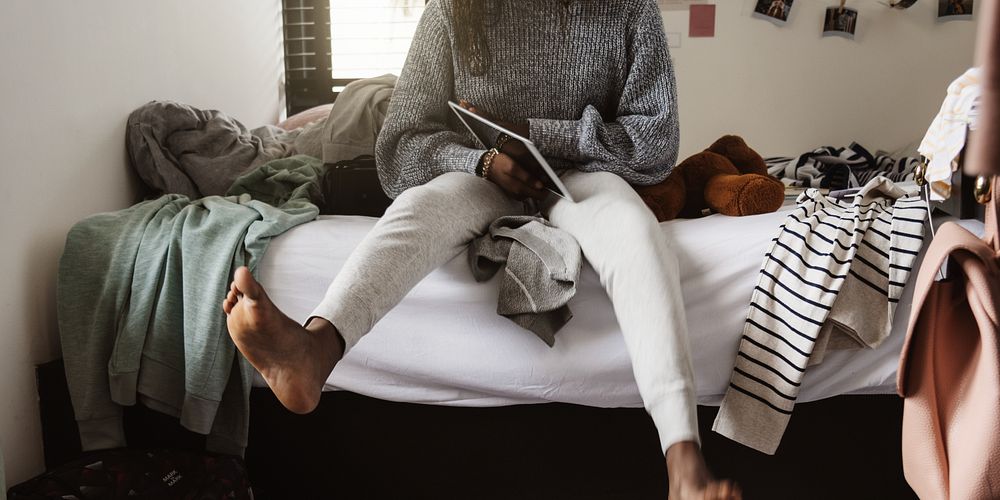 Person in cozy sweater and sweatpants sits on bed with tablet. Casual, relaxed setting with clothes and teddy bear.…