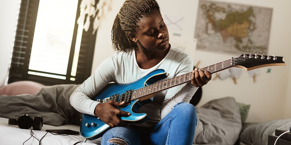 A black woman playing electric guitar on bed, focused and relaxed. Guitar practice in a cozy room, with a map on the wall.…