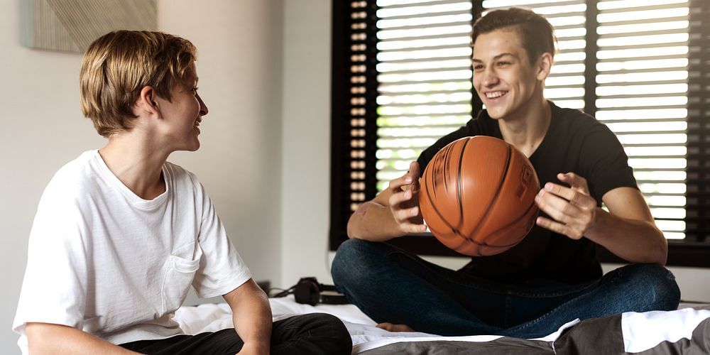 Two young males sitting indoors, one holding a basketball. They are casually dressed, smiling, and appear to be enjoying a…