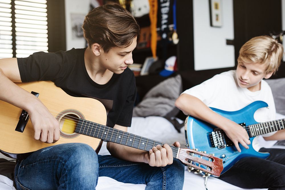 Two boys playing guitars in a cozy room. One with an acoustic guitar, the other with an electric guitar. Casual setting…