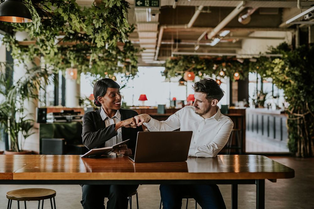 Two people in a modern office, engaged in a friendly fist bump. They sit at a wooden table with a laptop, surrounded by…