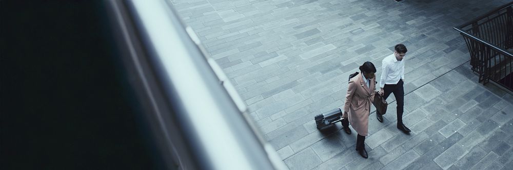 Two people walking on a paved path, one with a suitcase. Overhead view of a man and woman, dressed in business attire…