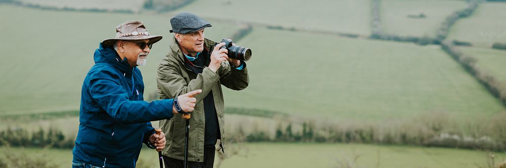 Two older men outdoors, one with a camera, enjoying nature. Both wear hats and jackets, standing in a green field, capturing…