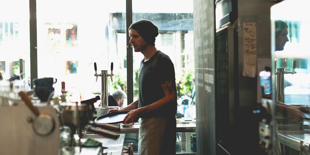 A barista in a beanie works in a cafe, preparing drinks. The cafe setting is cozy, with coffee equipment visible. The…