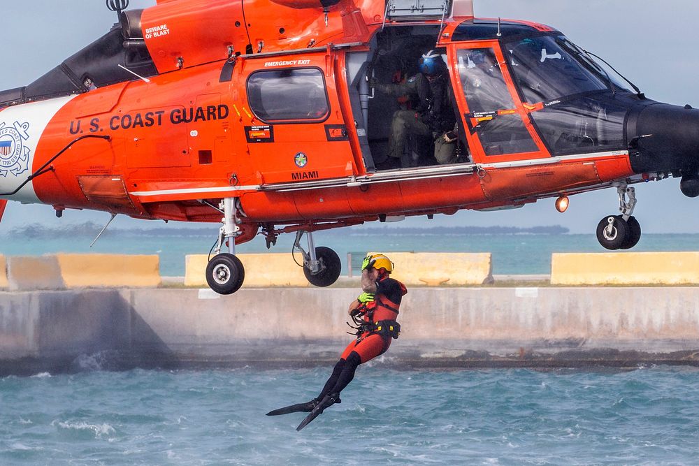 A U.S. Coast Guard rescue swimmer assigned to Air Station Miami jumps out of a, MH-65D Dolphin helicopter to simulate…