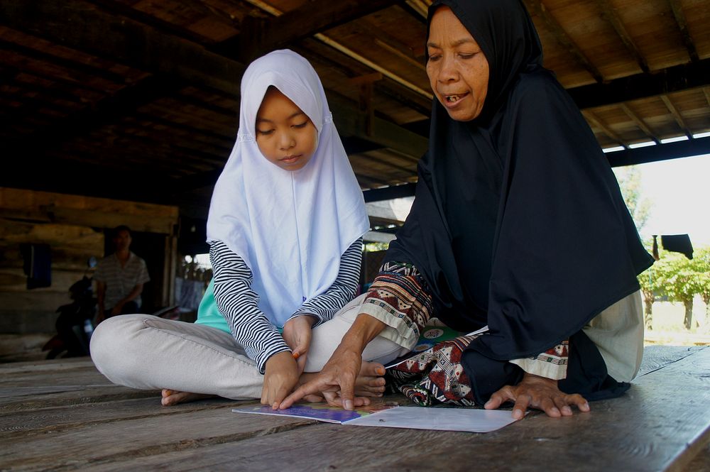 Membaca Bersama Ibu. Aisyah mengajar ibunya belajar membaca buku cerita. Photo by USAID Indonesia. Original public domain…