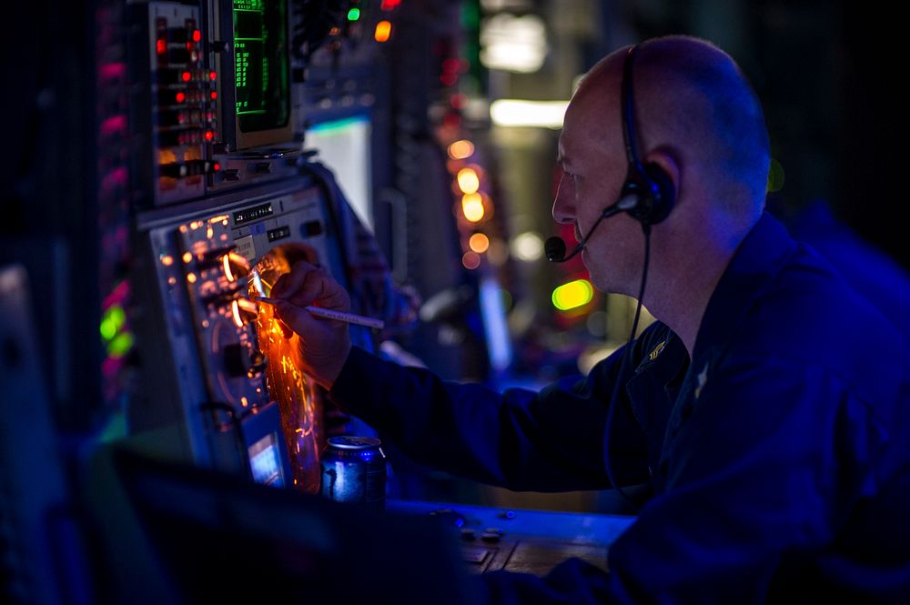 U.S. Navy Operations Specialist 1st Class Dale Schuler stands watch in the combat information center aboard the guided…