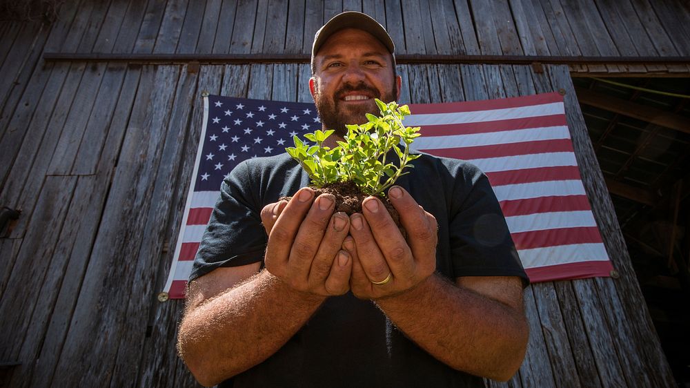 Holding an oregano seedling and soil, Calvin Riggleman is a Marine, served in Iraq, and now he serves his community farm…