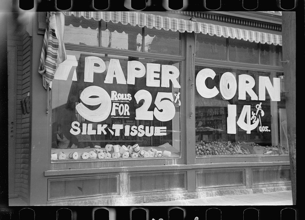 A sign in grocery store window "PAPER - 9 rolls for 25 cents CORN 14 1/2 cents," Manchester, New Hampshire. Sourced from the…