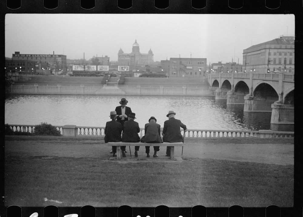 Men sitting on bench along Des Moines River. State capitol in background. Des Moines, Iowa. Sourced from the Library of…