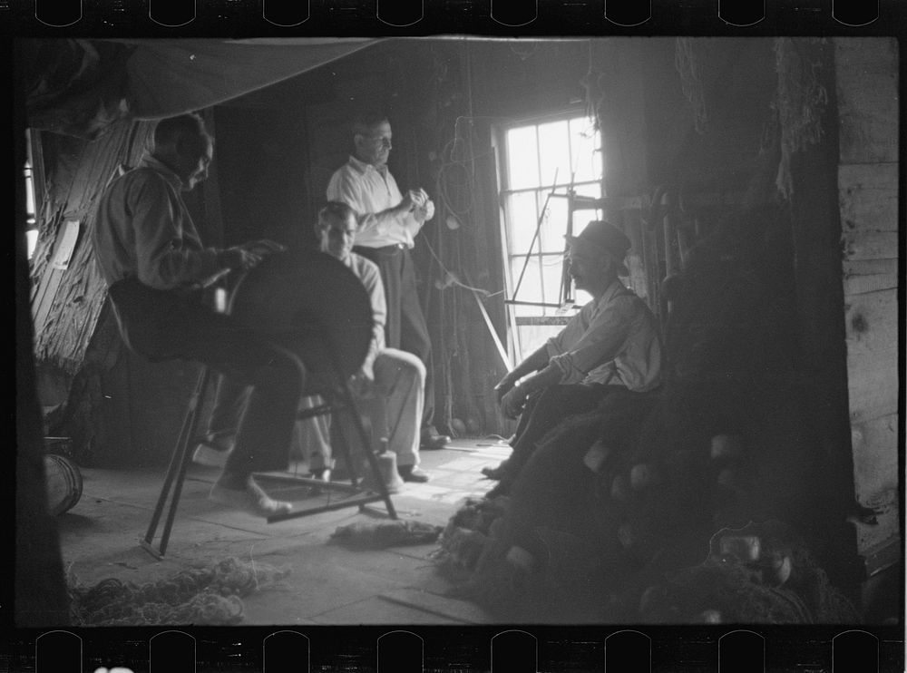 [Untitled photo, possibly related to: Fishermen in repair shack repairing nets and sharpening tools. The twine of these nets…